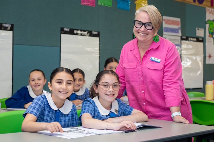A female teacher standing at the end of the desk with 2 girl students. 3 girl students are sitting behind